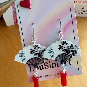 Op de afbeelding: Witte en zwarte waaier-vormige oorbellen met een bamboe-ontwerp en rode kraalaccenten. De oorbellen hebben zilverkleurige haken en worden gepresenteerd op een kaart met de tekst "LouSimArt.Etsy.com".