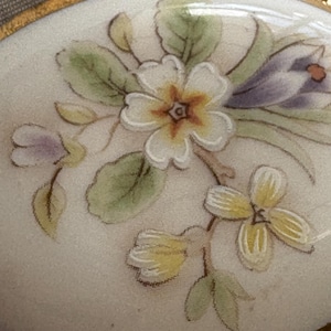 Może przedstawiać: Owalne pudełko na bibeloty z porcelany ze złotą obw&oacute;dką. Wieczko zdobi ręcznie malowany kwiatowy wz&oacute;r z białymi, ż&oacute;łtymi i fioletowymi kwiatami oraz zielonymi liśćmi. Pudełko ma błyszczące wykończenie.