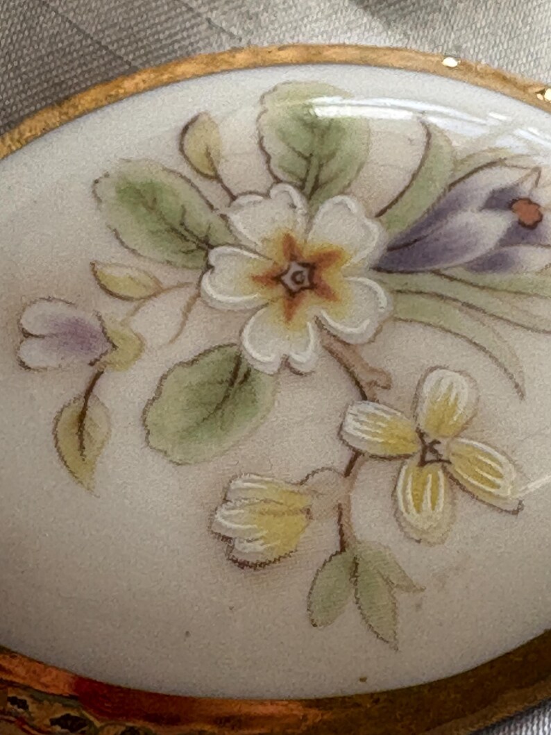 Może przedstawiać: Owalne pudełko na bibeloty z porcelany ze złotą obw&oacute;dką. Wieczko zdobi ręcznie malowany kwiatowy wz&oacute;r z białymi, ż&oacute;łtymi i fioletowymi kwiatami oraz zielonymi liśćmi. Pudełko ma błyszczące wykończenie.