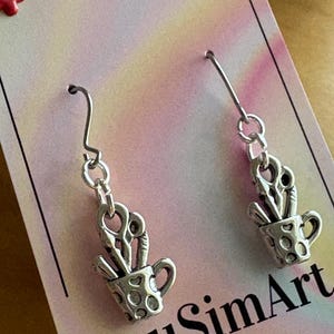 May include: Silver-toned earrings featuring a small cup with scissors inside. The cup has a dotted pattern. The earrings hang from a simple hook. The background is a pink and yellow gradient with the text "LousimArt.Etsy.com" and "LousimArt".