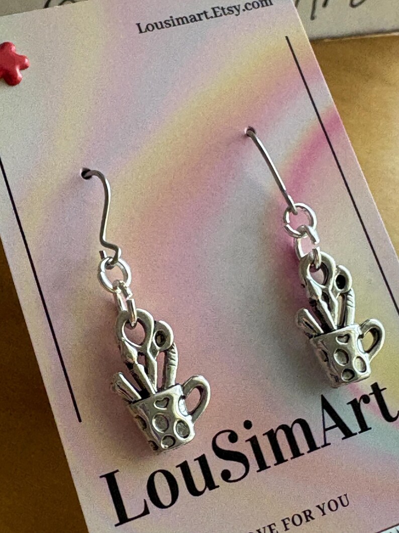 May include: Silver-toned earrings featuring a small cup with scissors inside. The cup has a dotted pattern. The earrings hang from a simple hook. The background is a pink and yellow gradient with the text "LousimArt.Etsy.com" and "LousimArt".