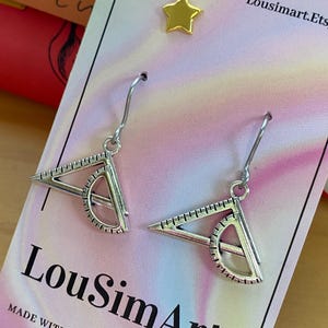 Peut inclure: Boucles d'oreilles en argent en forme de rapporteurs avec un petit cercle au centre. Les boucles d'oreilles sont sur une carte blanche avec le texte "LouSimArt" et "MADE WITH LOVE FOR YOU".