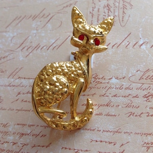 May include: Gold-toned cat brooch with red rhinestone eyes. The vintage pin features a textured body and a detailed design. A stylish accessory for cat lovers.