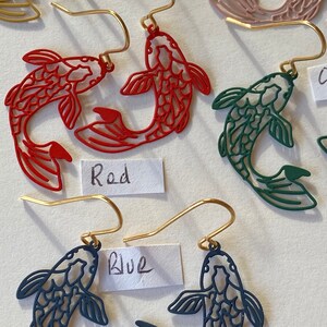 K&ouml;nnte beinhalten: Goldene Ohrh&auml;nger mit einem Paar Koi-Fischen in Rot, Blau und Gr&uuml;n. Die Fische sind aufwendig detailliert und haben ein filigranes, durchbrochenes Design. Die Ohrringe sind mit "Rot" und "Blau" beschriftet.