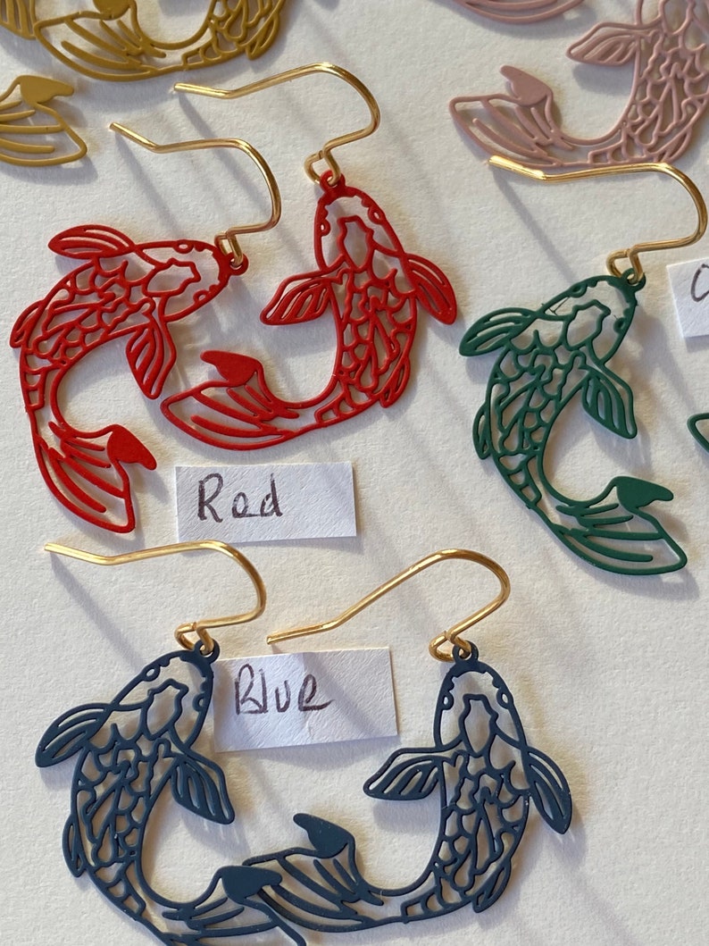 K&ouml;nnte beinhalten: Goldene Ohrh&auml;nger mit einem Paar Koi-Fischen in Rot, Blau und Gr&uuml;n. Die Fische sind aufwendig detailliert und haben ein filigranes, durchbrochenes Design. Die Ohrringe sind mit "Rot" und "Blau" beschriftet.