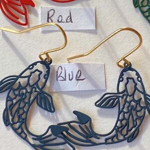 K&ouml;nnte beinhalten: Ein Paar blaue fischf&ouml;rmige Ohrringe mit goldenen Haken. Die Ohrringe sind aus Metall und haben ein filigranes, kompliziertes Design. Das Wort "Blau" steht auf einem St&uuml;ck Papier hinter den Ohrringen.