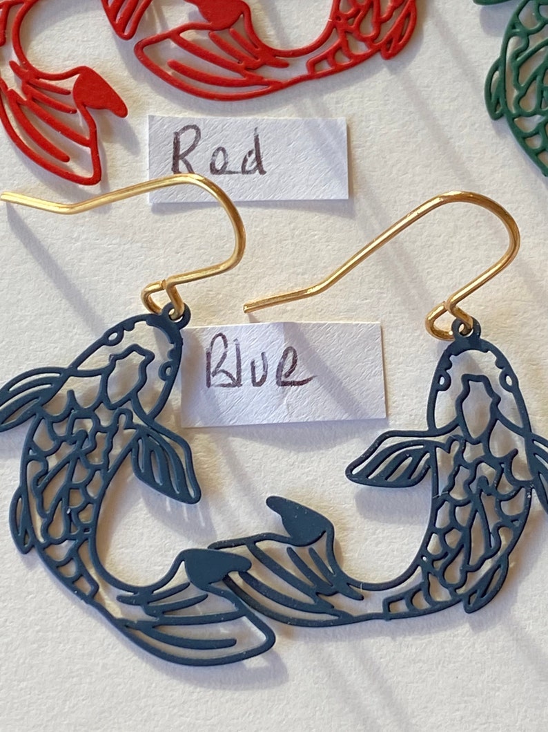 K&ouml;nnte beinhalten: Ein Paar blaue fischf&ouml;rmige Ohrringe mit goldenen Haken. Die Ohrringe sind aus Metall und haben ein filigranes, kompliziertes Design. Das Wort "Blau" steht auf einem St&uuml;ck Papier hinter den Ohrringen.