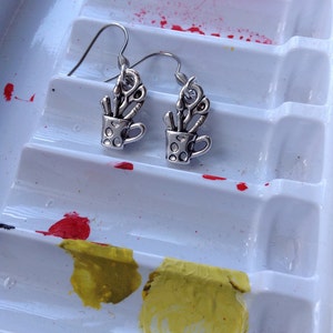 May include: Silver-toned earrings featuring a small cup design filled with scissors and other tools. The earrings are set against a white paint palette with red and yellow paint splotches, suggesting an art or craft theme.