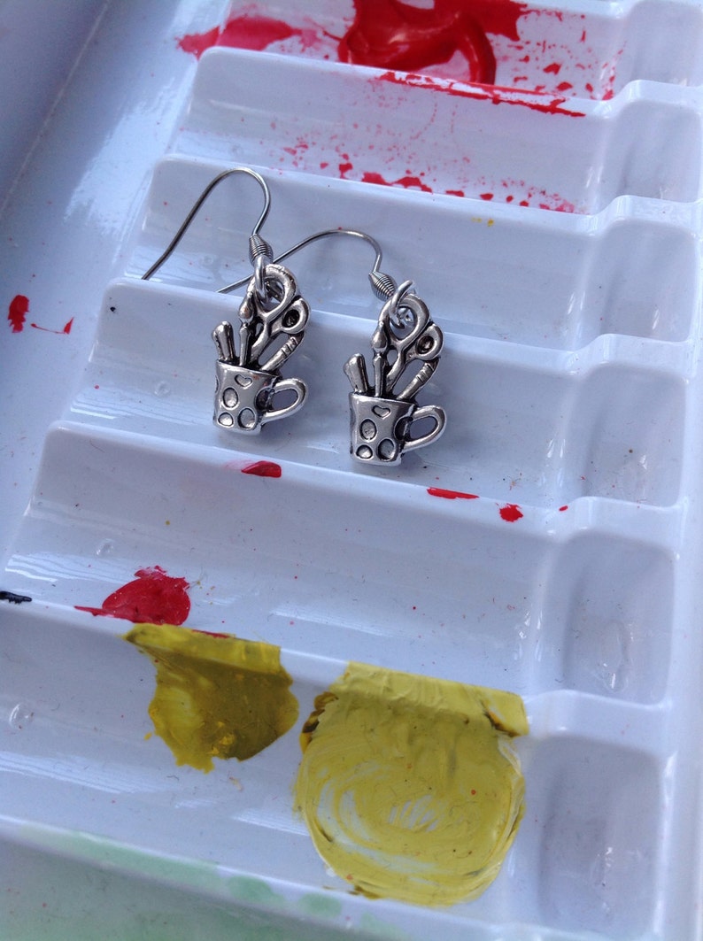 May include: Silver-toned earrings featuring a small cup design filled with scissors and other tools. The earrings are set against a white paint palette with red and yellow paint splotches, suggesting an art or craft theme.