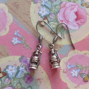 May include: A pair of silver dangle earrings featuring a lantern design. The earrings have a hook closure and are set against a floral patterned background.