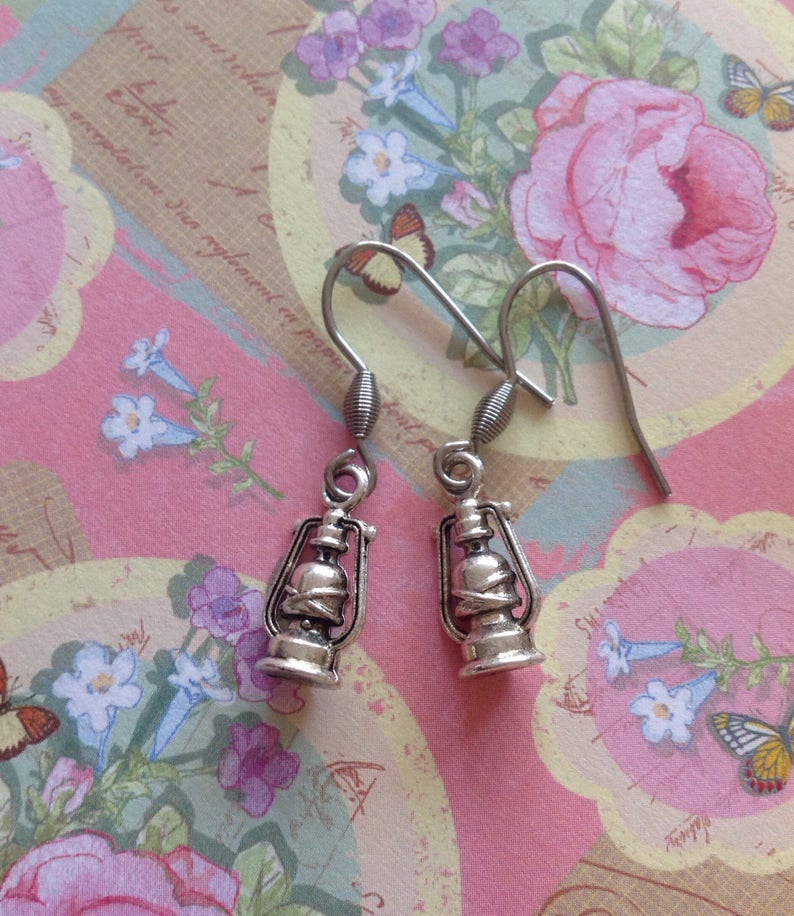 May include: A pair of silver dangle earrings featuring a lantern design. The earrings have a hook closure and are set against a floral patterned background.