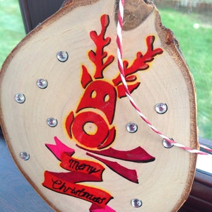 May include: A wooden Christmas ornament with a red reindeer design and the words "Merry Christmas" in pink lettering. The ornament is decorated with rhinestones.