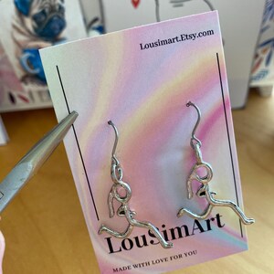 May include: Silver running figure earrings displayed on a pastel gradient card. The card features the text "LousimArt.etsy.com" and "MADE WITH LOVE FOR YOU". Pug-themed cards are visible in the background.