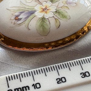 Może przedstawiać: Owalna broszka porcelanowa z ręcznie malowanym kwiatowym wzorem. Broszka ma kremową podstawę i złoty, teksturowany brzeg. Kwiatowy wz&oacute;r zawiera białe, ż&oacute;łte, fioletowe i zielone elementy. Broszka ma około 2,5 cm długości.