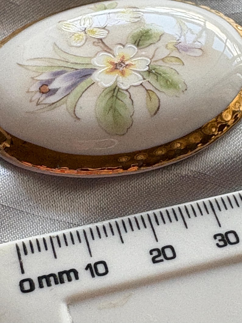 Może przedstawiać: Owalna broszka porcelanowa z ręcznie malowanym kwiatowym wzorem. Broszka ma kremową podstawę i złoty, teksturowany brzeg. Kwiatowy wz&oacute;r zawiera białe, ż&oacute;łte, fioletowe i zielone elementy. Broszka ma około 2,5 cm długości.