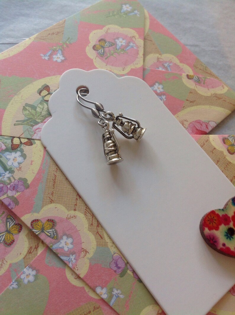 May include: Silver dangle earrings with a lantern charm. The earrings are displayed on a white tag with a pink and green floral pattern.
