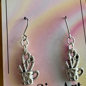 May include: Silver-toned earrings featuring a small cup charm filled with art supplies, including scissors and paintbrushes. The cup has a handle and a dotted pattern. The earrings hang from silver hooks. The text "LouSimArt" is visible.