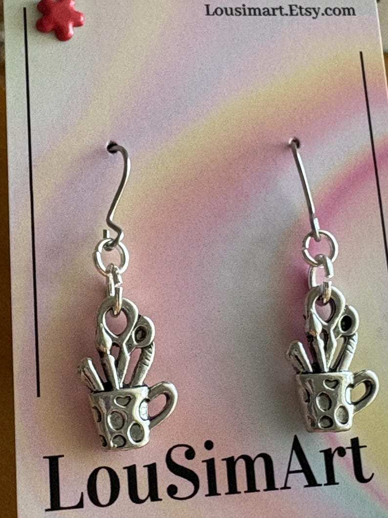 May include: Silver-toned earrings featuring a small cup charm filled with art supplies, including scissors and paintbrushes. The cup has a handle and a dotted pattern. The earrings hang from silver hooks. The text "LouSimArt" is visible.