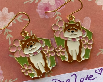 May include: A pair of gold-colored earrings featuring a Shiba Inu dog design. The dog is brown, white, and black, wearing a pink scarf and surrounded by pink flowers on a green background. The text "Husky Dog Love" is written on a white label.