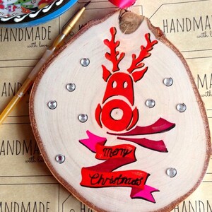 May include: A wooden Christmas ornament with a red reindeer and a pink and red banner that says "Merry Christmas". The ornament is decorated with silver glitter and rhinestones.