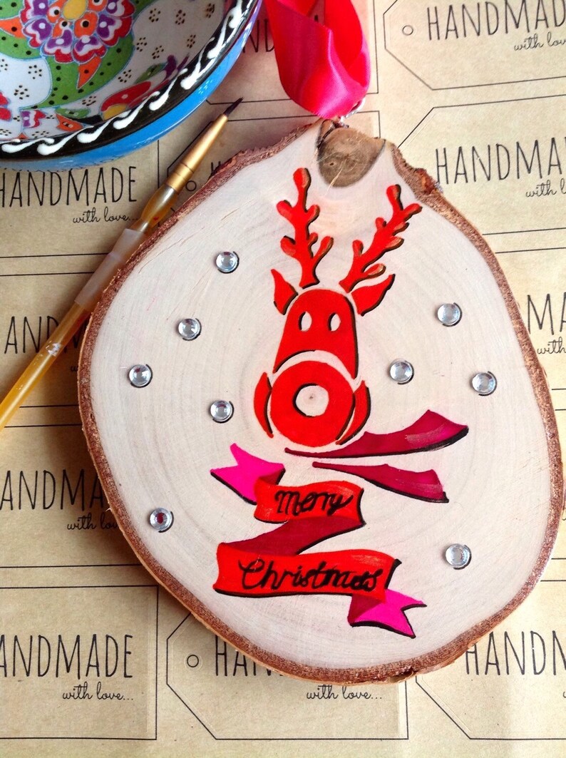 May include: A wooden Christmas ornament with a red reindeer and a pink and red banner that says "Merry Christmas". The ornament is decorated with silver glitter and rhinestones.