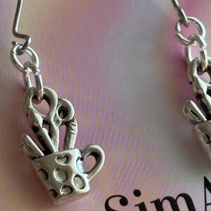 May include: Silver-toned earrings featuring a small cup design filled with art supplies. The cup has a handle and dotted pattern. The earrings hang from a simple hook and chain. The background is a soft pink and white.