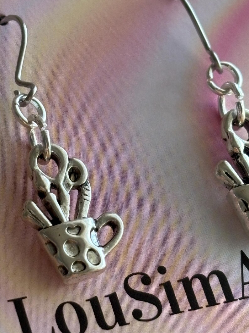 May include: Silver-toned earrings featuring a small cup design filled with art supplies. The cup has a handle and dotted pattern. The earrings hang from a simple hook and chain. The background is a soft pink and white.
