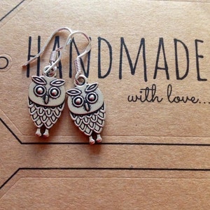 Pode incluir: Um par de brincos de prata em forma de coruja com fecho de gancho. Os brincos est&atilde;o em um cart&atilde;o marrom com o texto "HANDMADE with love..."