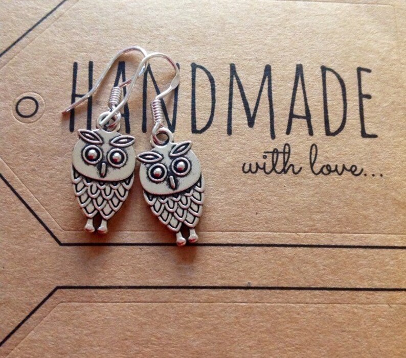 Pode incluir: Um par de brincos de prata em forma de coruja com fecho de gancho. Os brincos est&atilde;o em um cart&atilde;o marrom com o texto "HANDMADE with love..."