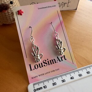 May include: Silver-toned earrings featuring a small cup design filled with scissors and other tools. The earrings hang from a silver hook. The product card has the text "LouSimArt" and "Made with love for you".
