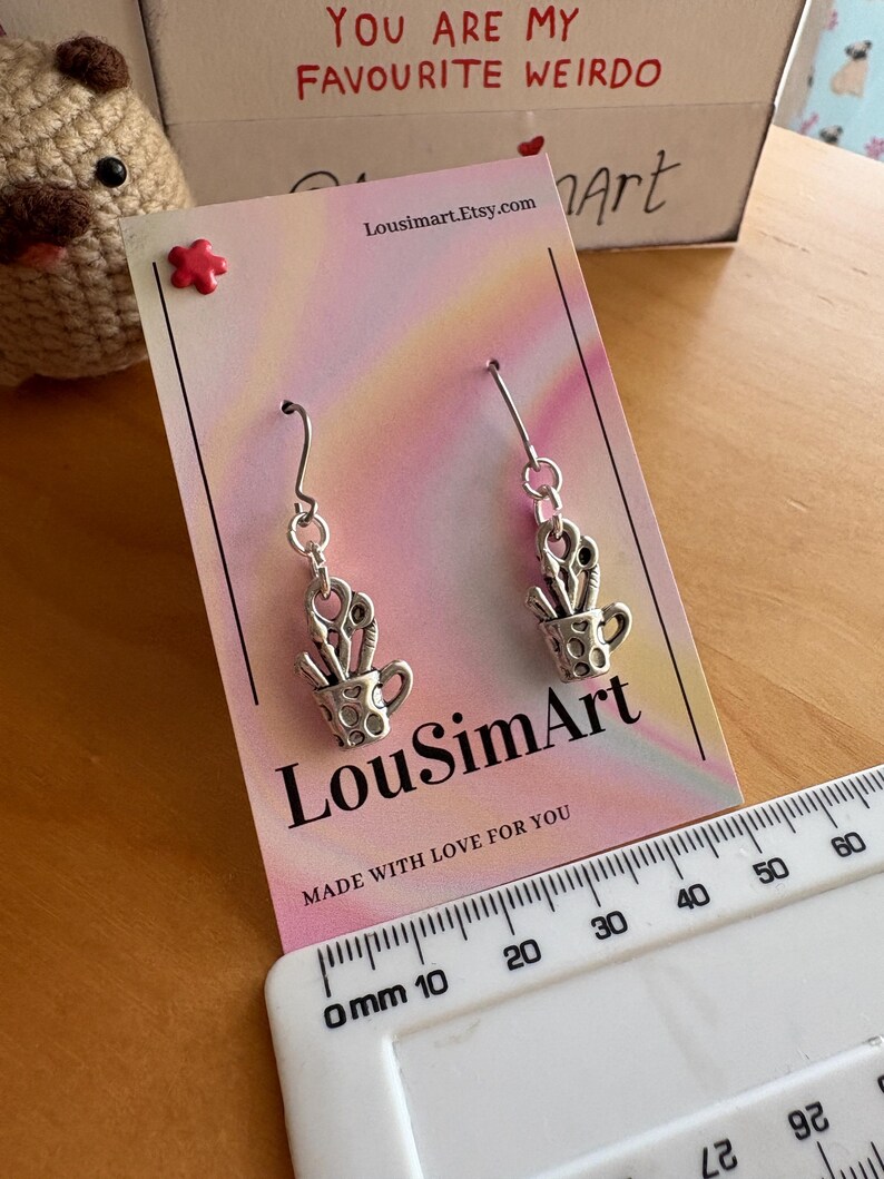May include: Silver-toned earrings featuring a small cup design filled with scissors and other tools. The earrings hang from a silver hook. The product card has the text "LouSimArt" and "Made with love for you".