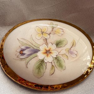 Może przedstawiać: Owalna broszka porcelanowa ze złotą obw&oacute;dką. Broszka przedstawia ręcznie malowany kwiatowy wz&oacute;r z białymi, ż&oacute;łtymi i fioletowymi kwiatami oraz zielonymi liśćmi. Tło jest kremowo-białe.