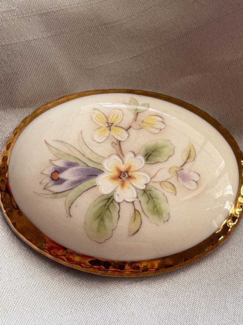 Może przedstawiać: Owalna broszka porcelanowa ze złotą obw&oacute;dką. Broszka przedstawia ręcznie malowany kwiatowy wz&oacute;r z białymi, ż&oacute;łtymi i fioletowymi kwiatami oraz zielonymi liśćmi. Tło jest kremowo-białe.