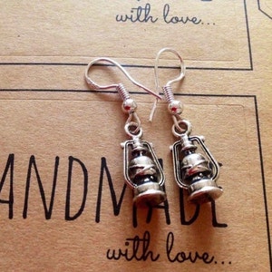 May include: Silver dangle earrings with a lantern charm. The earrings are handmade and the charms are detailed with a silver finish. The earrings are on a brown background with the text "Handmade with love..."