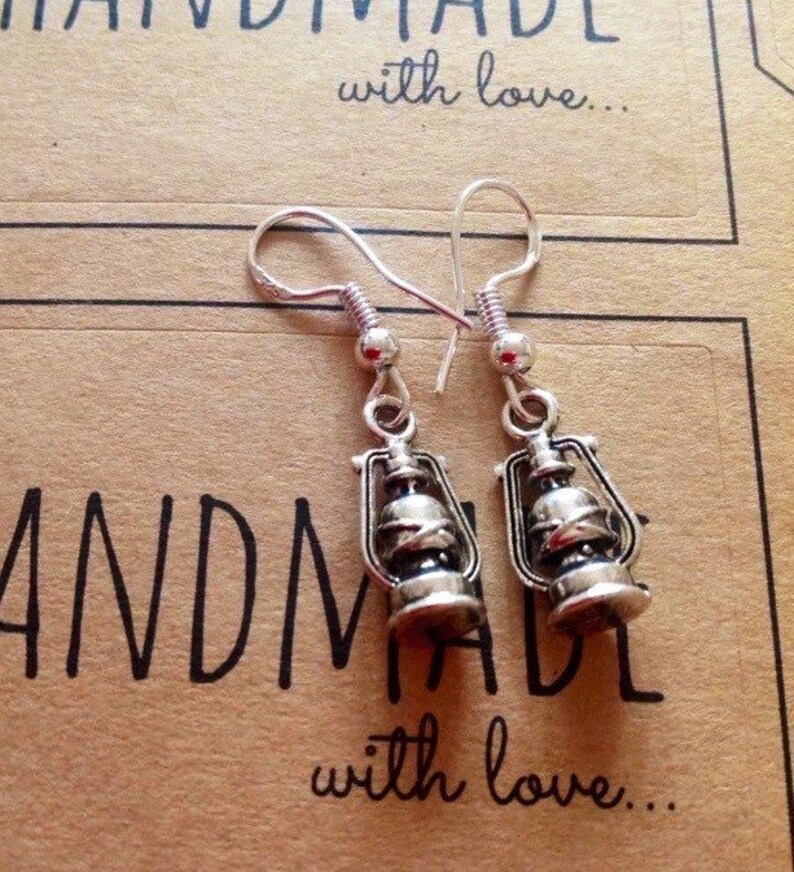 May include: Silver dangle earrings with a lantern charm. The earrings are handmade and the charms are detailed with a silver finish. The earrings are on a brown background with the text "Handmade with love..."