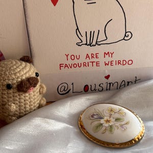 Może przedstawiać: Owalna porcelanowa broszka ze złotą obw&oacute;dką, z kwiatowym wzorem w pastelowych kolorach. Widoczny jest r&oacute;wnież szydełkowy mops i kartka z ilustracją mopsa i napisem "YOU ARE MY FAVOURITE WEIRDO".