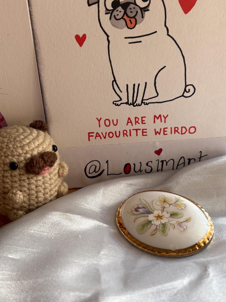 Może przedstawiać: Owalna porcelanowa broszka ze złotą obw&oacute;dką, z kwiatowym wzorem w pastelowych kolorach. Widoczny jest r&oacute;wnież szydełkowy mops i kartka z ilustracją mopsa i napisem "YOU ARE MY FAVOURITE WEIRDO".