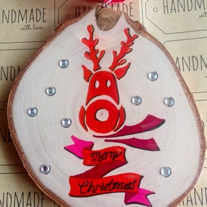 May include: A wooden Christmas ornament with a red reindeer painted on it. The reindeer has a red nose and antlers. The ornament also has a red and pink ribbon with the words "merry Christmas" written on it.