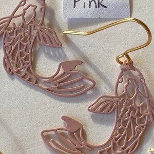 K&ouml;nnte beinhalten: Ein Paar rosa fischf&ouml;rmige Ohrringe mit filigranen Ausschnitten. Die Ohrringe sind aus Metall und haben goldene Haken.