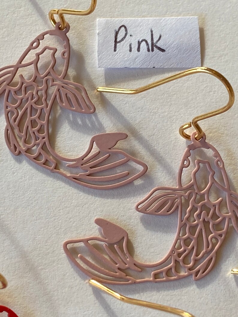 K&ouml;nnte beinhalten: Ein Paar rosa fischf&ouml;rmige Ohrringe mit filigranen Ausschnitten. Die Ohrringe sind aus Metall und haben goldene Haken.