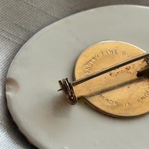 Może przedstawiać: Owalna broszka z białej porcelany ze złotym metalowym tyłem. Szpilka ma złote zapięcie i szpilkę. Z tyłu broszki znajduje się napis "Made in England".