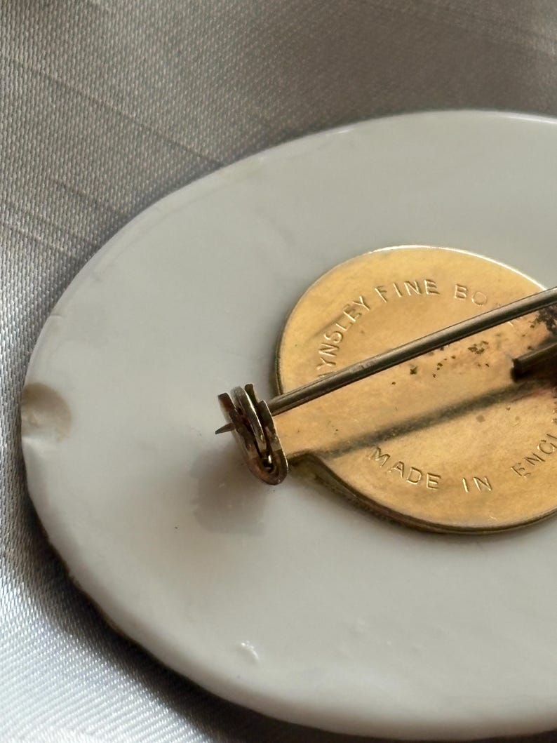 Może przedstawiać: Owalna broszka z białej porcelany ze złotym metalowym tyłem. Szpilka ma złote zapięcie i szpilkę. Z tyłu broszki znajduje się napis "Made in England".