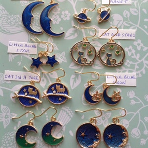 May include: A collection of 10 pairs of gold-tone earrings with blue and green enamel. The earrings feature various designs, including moons, stars, cats, and planets. The earrings are labeled with names like "Big Blue Moon", "Little Blue Planet", "Cat and Stars", "Cat in a Tree", "Little Blue Moon", "Blue Green Moon", and "Gold Moon and Star".