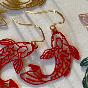 K&ouml;nnte beinhalten: Rote Metallohrringe in Form von zwei Koi-Fischen mit einem goldenen Haken. Das Wort "Rot" steht auf einem wei&szlig;en Papier unter den Ohrringen.