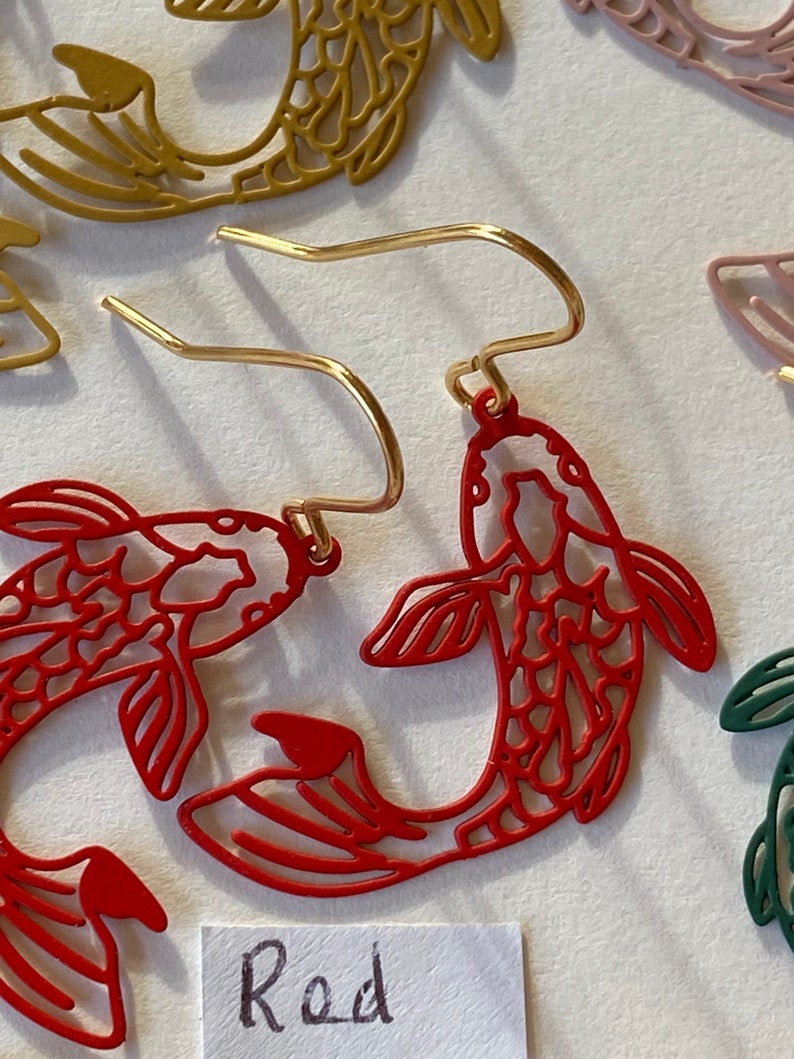 K&ouml;nnte beinhalten: Rote Metallohrringe in Form von zwei Koi-Fischen mit einem goldenen Haken. Das Wort "Rot" steht auf einem wei&szlig;en Papier unter den Ohrringen.