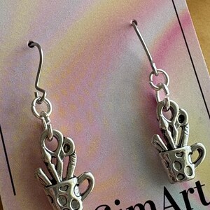 May include: Silver-toned earrings featuring a cup design filled with scissors and paintbrushes. The cup has a handle and is decorated with small, round cutouts. The earrings hang from silver-colored ear wires. The background is a colorful, pastel-toned card.