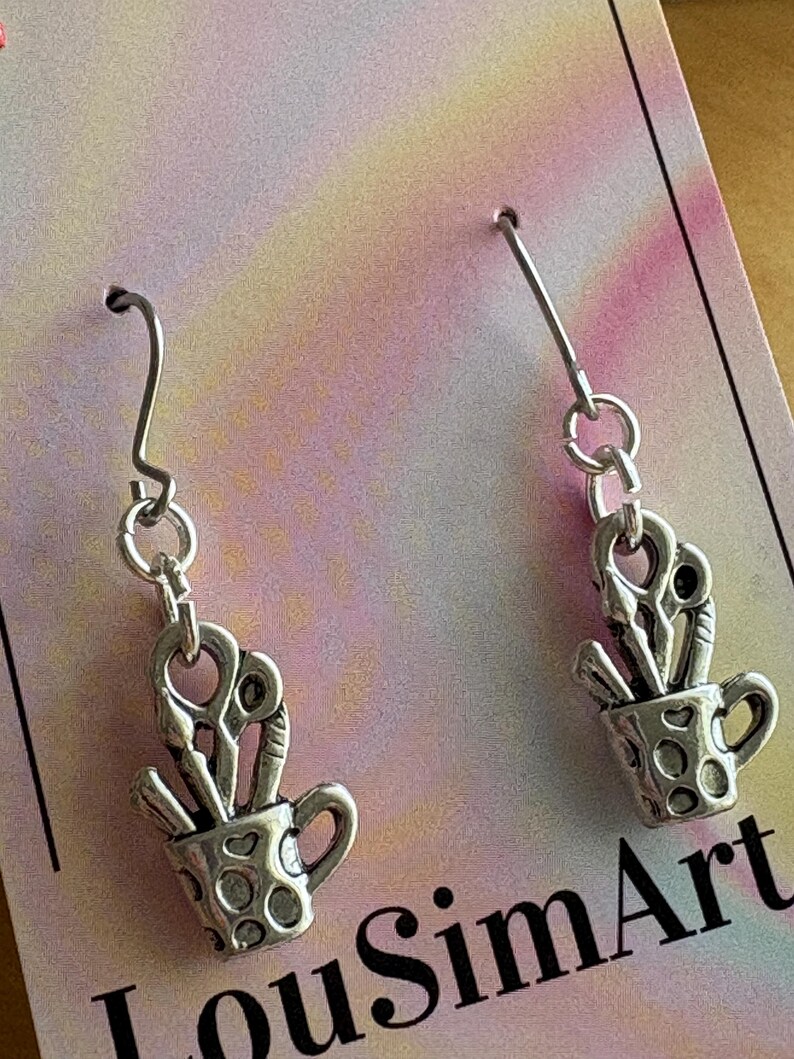 May include: Silver-toned earrings featuring a cup design filled with scissors and paintbrushes. The cup has a handle and is decorated with small, round cutouts. The earrings hang from silver-colored ear wires. The background is a colorful, pastel-toned card.