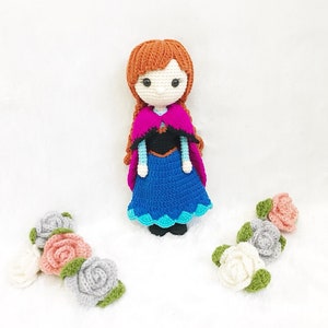 May include: A crocheted doll dressed as Anna from the movie Frozen. The doll has brown hair, a blue dress, a pink cape, and a black bodice. The doll is surrounded by crocheted flowers in shades of pink, white, and gray.