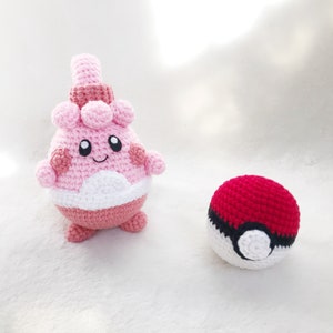 May include: Crocheted Clefairy and Pok&eacute; Ball. The Clefairy is pink, white, and dark pink with a smiling face. The Pok&eacute; Ball is red and white with a black stripe. Both are handmade and sit on a white surface.