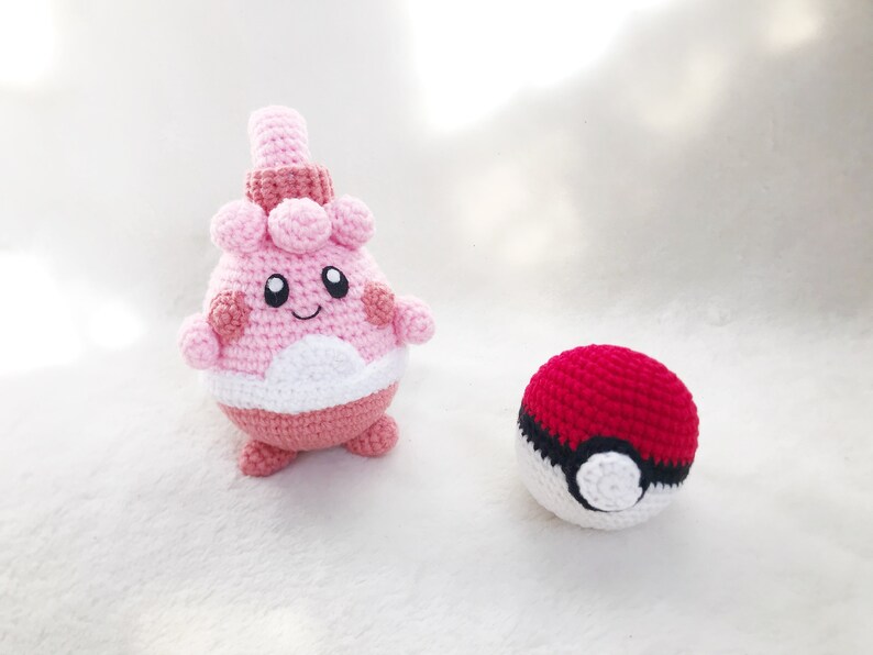May include: Crocheted Clefairy and Pok&eacute; Ball. The Clefairy is pink, white, and dark pink with a smiling face. The Pok&eacute; Ball is red and white with a black stripe. Both are handmade and sit on a white surface.
